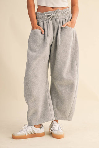 A person wearing light heather gray wide-leg sweatpants with an elastic drawstring waist and side pockets, paired with white sneakers, against a plain beige studio background.