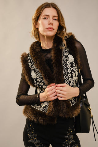 Woman wearing a black outfit with a fur-trimmed vest and patterned scarf against a neutral background