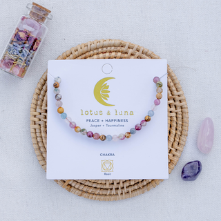 A beaded chakra bracelet by lotus & luna, featuring multicolored gemstone beads, displayed on its white backing card. The card is placed on a round woven straw mat, with a small glass bottle of dried flowers and two polished crystals nearby on a textured white fabric background.