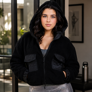A woman wearing a black hooded zip-up jacket with large front flap pockets and metallic silver leggings, standing indoors near large glass doors.