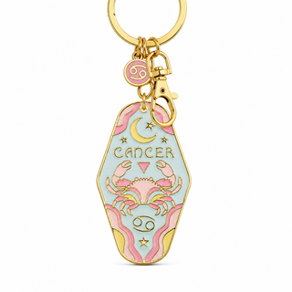A gold-tone metal keychain charm shaped like a coffin, featuring pastel pink and light blue enamel artwork of a crab, crescent moon, stars, and the word 'CANCER' with the Cancer zodiac symbol. A small round pink charm with a zodiac symbol hangs above the coffin.