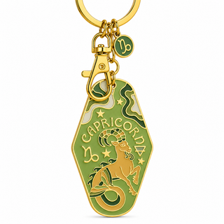 A gold-tone metal keychain featuring a green and gold Capricorn zodiac pendant with an engraved goat illustration and the word 'CAPRICORNIA'. The keychain includes a lobster clasp, a gold ring, and a small round charm with the Capricorn symbol.