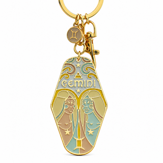A gold-tone Gemini zodiac keychain charm shaped like a hamsa hand, featuring light blue, beige, and brown enamel details with the word 'Gemini' and a small Gemini zodiac symbol charm above it, set against a plain white background.