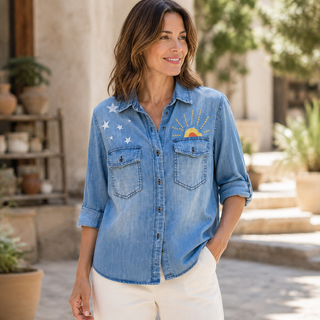 A woman wearing a light blue denim button-up shirt with embroidered white stars and a sun motif on the chest, paired with white pants, standing in a sunlit outdoor patio setting.