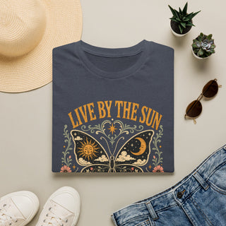 A folded navy graphic T-shirt is arranged in a styled flat lay on a light beige surface. The shirt features a large butterfly design with a sun and moon inside the wings, surrounded by floral details and the text 'LIVE BY THE SUN' in warm gold lettering.