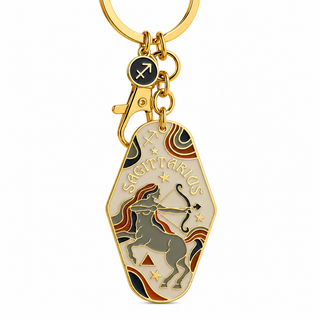 A gold-tone metal keychain featuring a coffin-shaped pendant with an engraved zodiac design for Sagittarius, including the word 'SAGITTARIUS', a bow and arrow, a horse, stars, and decorative wavy patterns. The pendant has a cream background with gold, brown, and red details, and is attached to a gold clasp and a small round charm with a zodiac symbol.