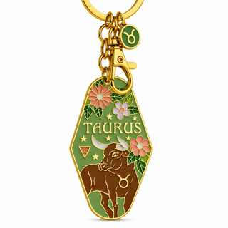 A gold-tone metal keychain featuring a green coffin-shaped pendant with the word 'TAURUS' and an illustration of a brown bull, pink flowers, and green leaves. A small green circular charm with a zodiac symbol hangs from the key ring on a white background.