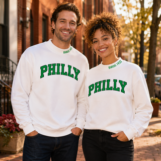 White man and light-skinned Black woman wearing matching white PHILLY mockneck sweatshirts with green BIRDS 1933 collar embroidery.