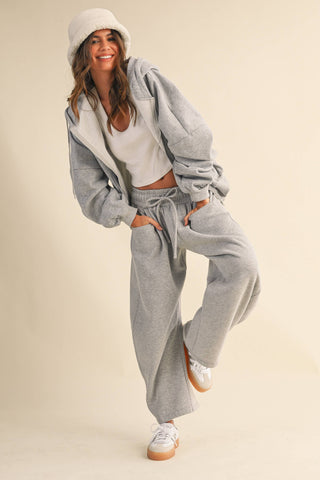 A woman modeling a light gray two-piece lounge set consisting of a zip-up hoodie and matching drawstring sweatpants, styled with a white crop top, white beanie, and white sneakers against a solid beige background.