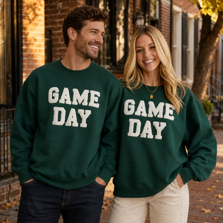 Man and woman wearing matching forest green GAME DAY terry-embroidered crewneck sweatshirts on a Philadelphia street.