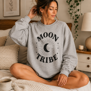 Woman wearing a gray sweatshirt with 'Moon Tribe' design, sitting on a bed.